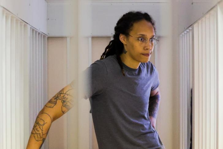 A tall Black woman wearing wire-framed glasses and a grey T-shirt stands in a prison sell