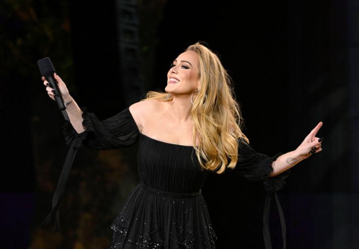 adele smiles on stage holding a wireless microphone in one hand wearing a black dress