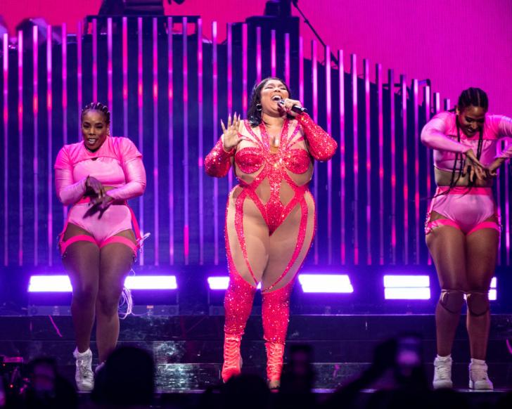 lizzo belts out a song into a microphone on stage wearing a sparkly pink costume
